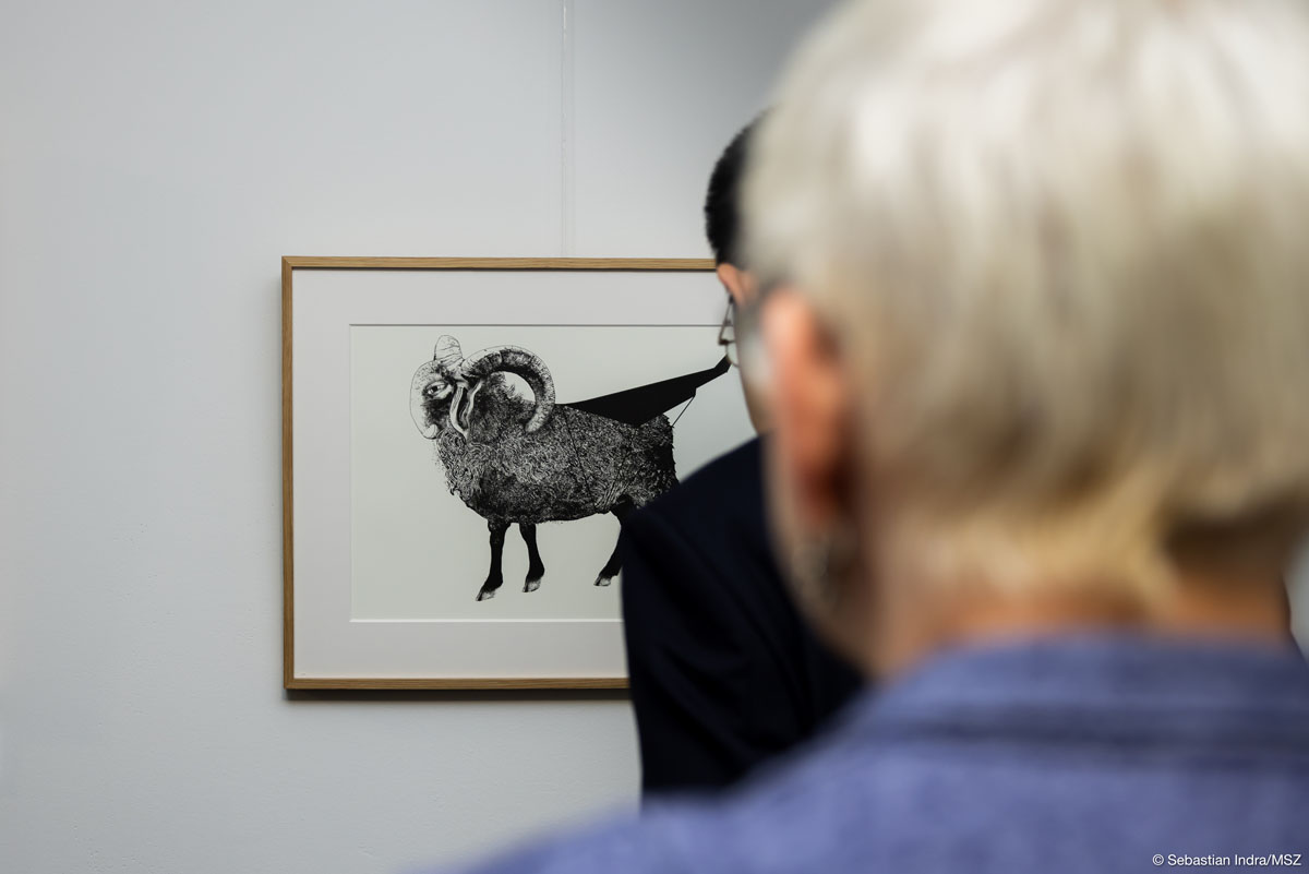 Na białej ścianie oprawiona w cienką drewnianą ramkę i białe passe-partout czarno-biała grafika z zwierzęcia kopytnego z dużymi zakręconymi rogami. Na pierwszym planie rozmazane sylwetki dwóch oglądających ją mężczyzn stojących jeden za drugim.