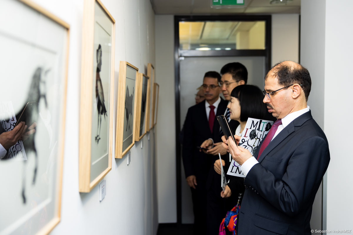 Pięcioro ludzi stoi w wąskim korytarzu i ogląda biało-czarne grafiki wiszące na białych ścianach. Mężczyzna na pierwszym planie w garniturze i okularach robi telefonem zdjęcie grafice lub sprawdza coś w telefonie.