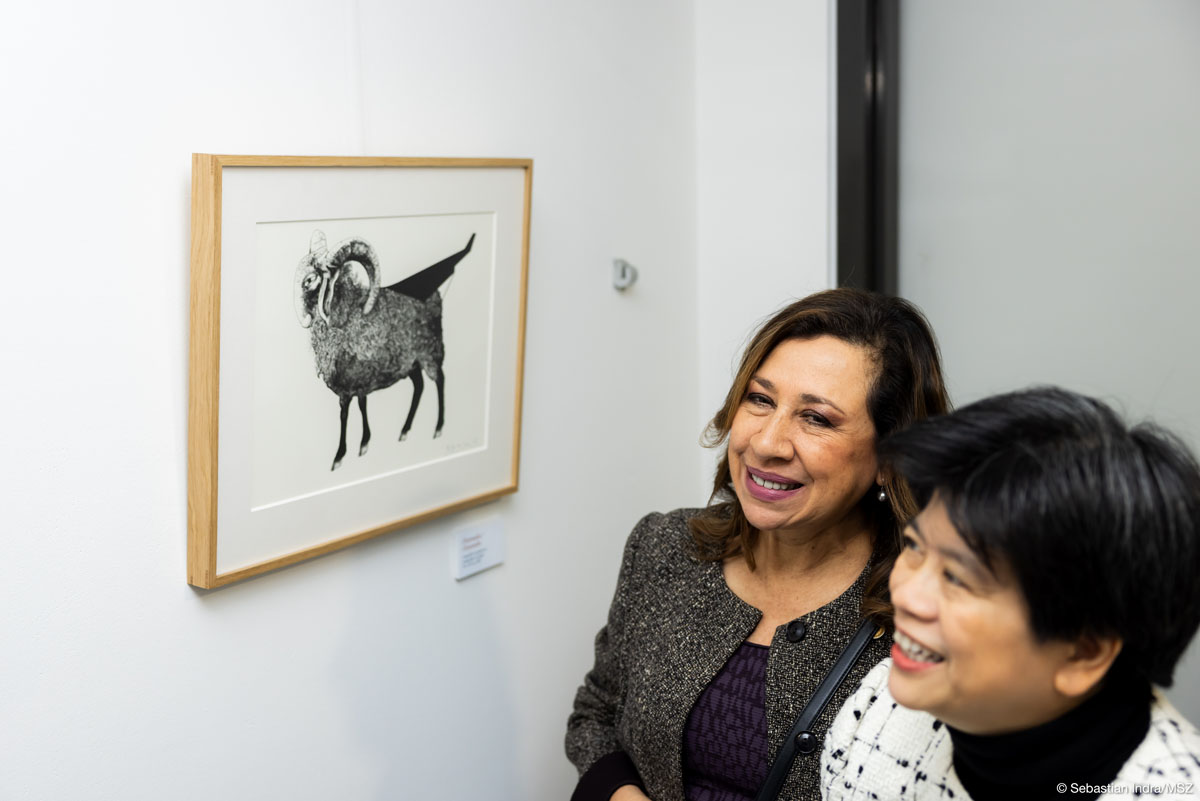 Dwie uśmiechające się kobiety patrzą na eksponowaną na białej ścianie czarno-białe grafikę, przedstawiającą zwierzę kopytne z zakręconymi rogami.