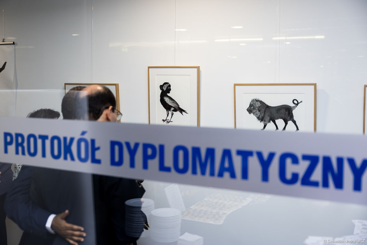Dwaj mężczyźni patrzą na eksponowane na białej ścianie czarno-białe grafiki. Stoją za szklaną ścianą z napisem „Protokół dyplomatyczny” widoczną na pierwszym planie.