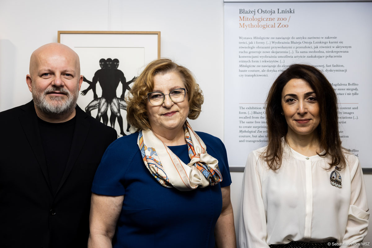 Troje ludzi: łysy mężczyzna z siwą brodą, w środku kobieta o blond półdługich włosach w okularach ubrana w granatową sukienkę z krótkimi rękawami i apaszką, po prawej kobieta z ciemnymi długimi włosami w białej bluzce pozują na tle czarno-białej grafiki i planszy z napisem: „Błażej Ostoja Lniski/ Mitologiczne zoo/ Mythological Zoo”.