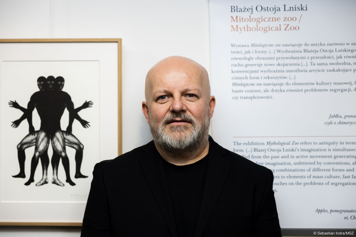 Łysy mężczyzna z siwą brodą ubrany na pozuje na tle czarno-białej grafiki i planszy z napisem: „Błażej Ostoja Lniski/ Mitologiczne zoo/ Mythological Zoo”.