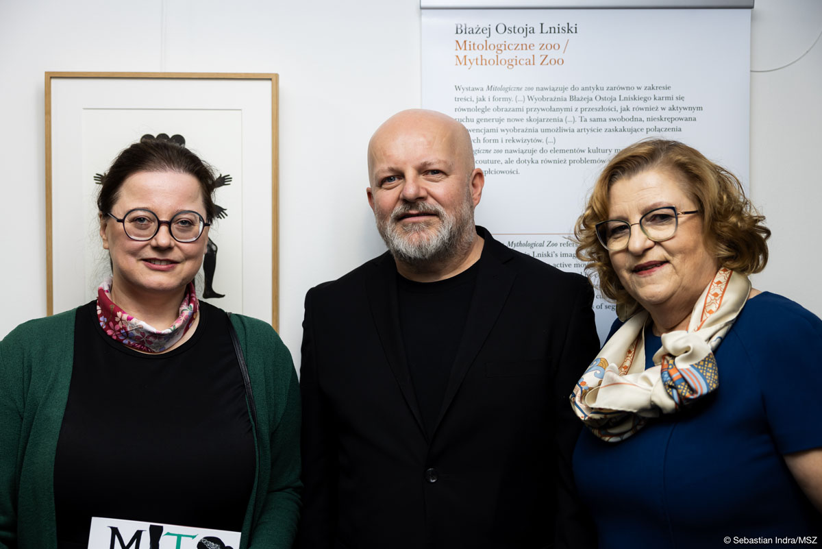 Troje ludzi: kobieta z gładko zaczesanymi do tyłu ciemnymi włosami w okularach, w środku łysy mężczyzna z siwą brodą, po prawej kobieta o blond półdługich włosach w okularach ubrana w granatową sukienkę z krótkimi rękawami i apaszką pozują na tle czarno-białej grafiki i planszy z napisem: „Błażej Ostoja Lniski/ Mitologiczne zoo/ Mythological Zoo”.