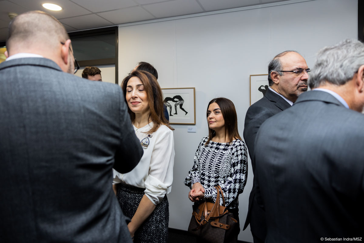 Kilkoro ludzi stoi w pomieszczeniu, w którym na białych ścianach wiszą czarno-białe grafiki. Ludzie tworzą grupy, na środku stoi kobieta z ciemnymi długimi włosami w czarno-białej bluzce z torebką przewieszoną na lewym przedramieniu. Patrzy w stronę jednego mężczyzny.