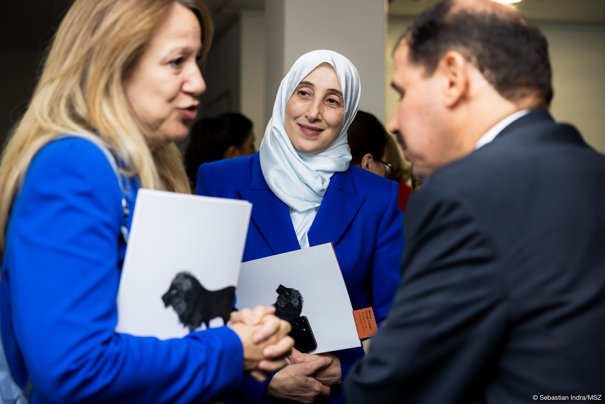 Na pierwszym planie nieco rozmazana para – kobieta o blond długich włosach w granatowej marynarce zwraca się do mężczyzny z wąsami w granatowym garniturze. Pośrodku pomiędzy nimi wyraźnie widoczna kobieta w białym hidżabie i granatowej marynarce spogląda na mówiącą kobietę lekko się uśmiechając. Obie kobiety trzymają w rękach katalog z wystawy.