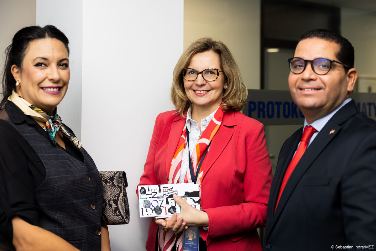 Trzy osoby pozują: po lewej kobieta z upiętymi do tyłu ciemnymi włosami w ciemnej kamizelce i kolorowej apaszce z torebką z wężowej skóry pod prawą pachą, w środku kobieta w okularach z blond półdługimi włosami w czerwonej marynarce i luźno przerzuconej kolorowej apaszce z identyfikatorem na szyi i katalogiem z wystawy w rękach, po prawej ciemnowłosy mężczyzna w okularach w czarnej marynarce i czerwonym krawacie. Wszyscy troje uśmiechają się.