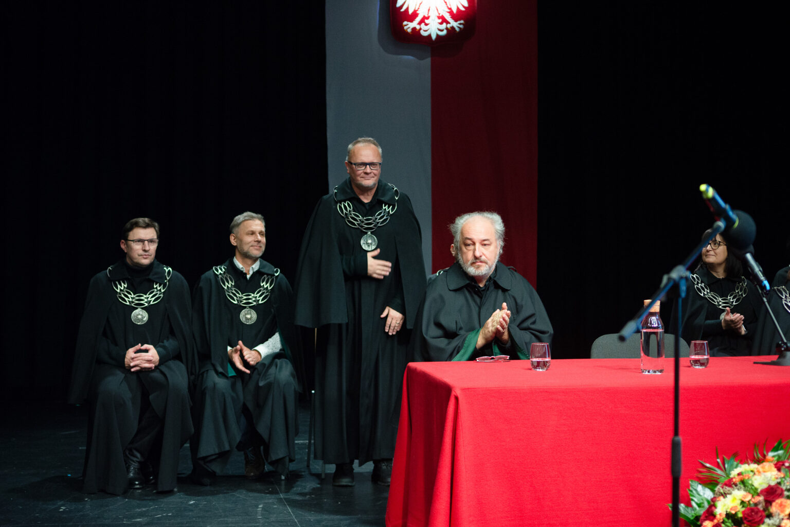 mężczyźni w strojach reprezentacyjnych ASP w Warszawie przy czerwonym stole, godło w tle