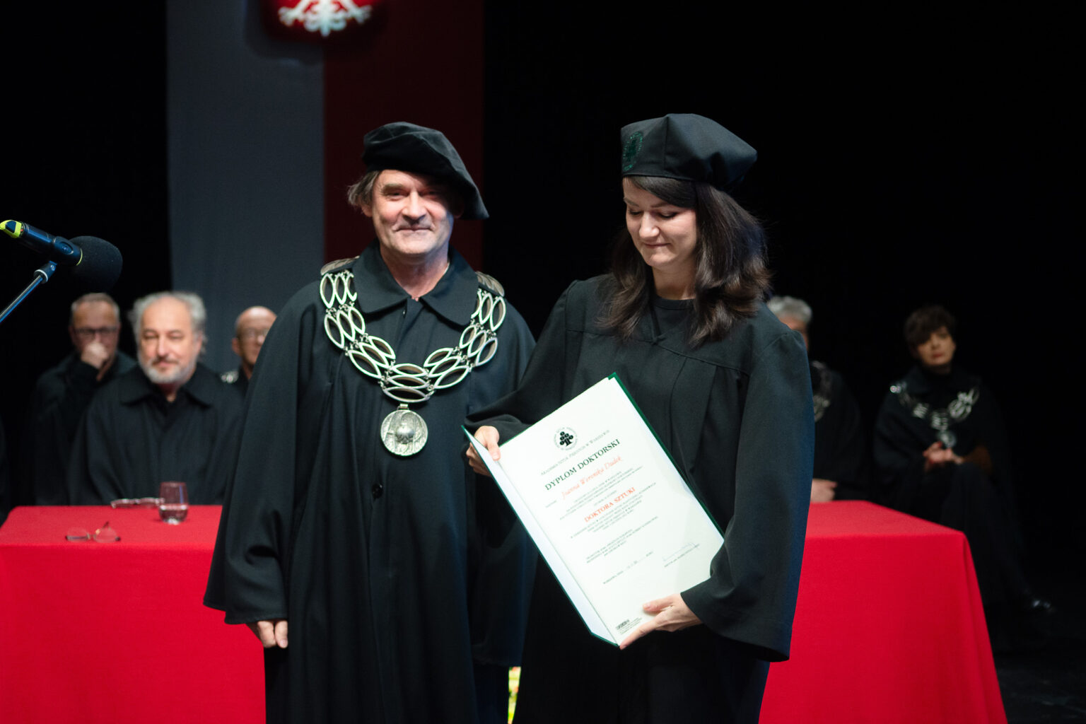 mężczyzna i kobieta w strojach reprezentacyjnym ASP w Warszawie, kobieta prezentuje dyplom