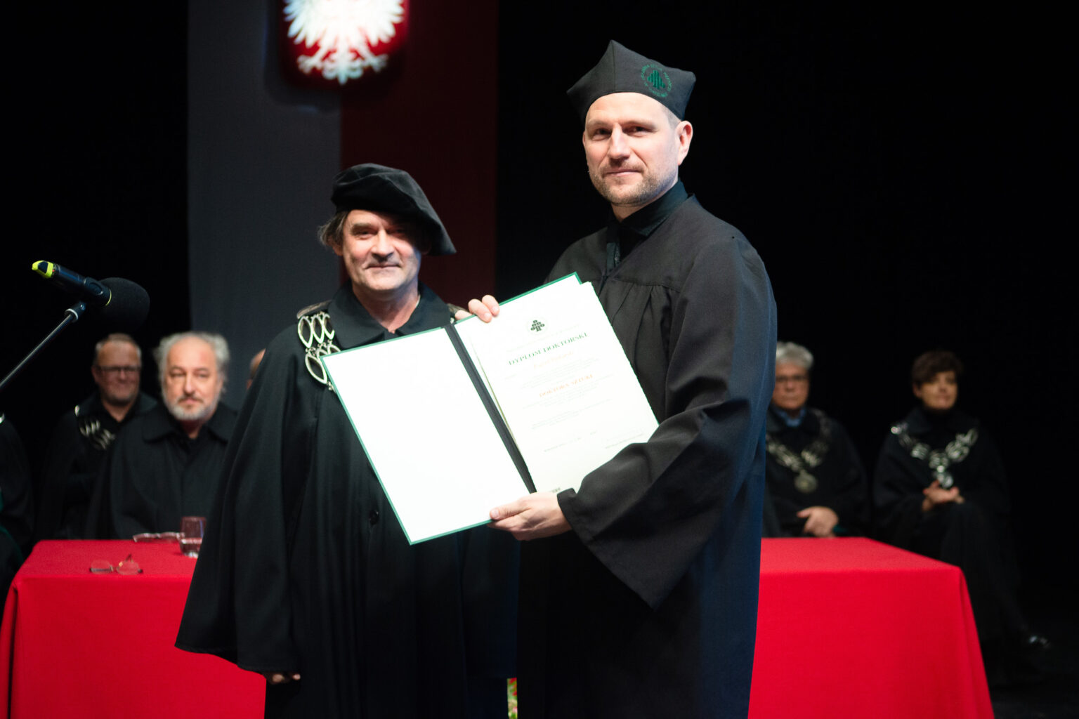 dwaj mężczyźni w strojach reprezentacyjnym ASP w Warszawie, jeden z nich prezentuje dyplom