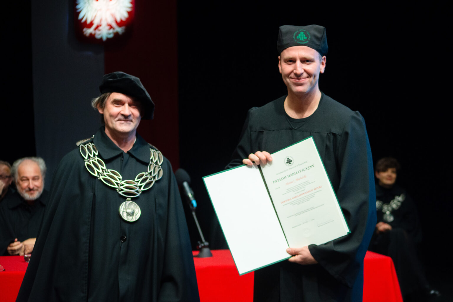 dwaj mężczyźni w strojach reprezentacyjnym ASP w Warszawie, jeden z nich prezentuje dyplom