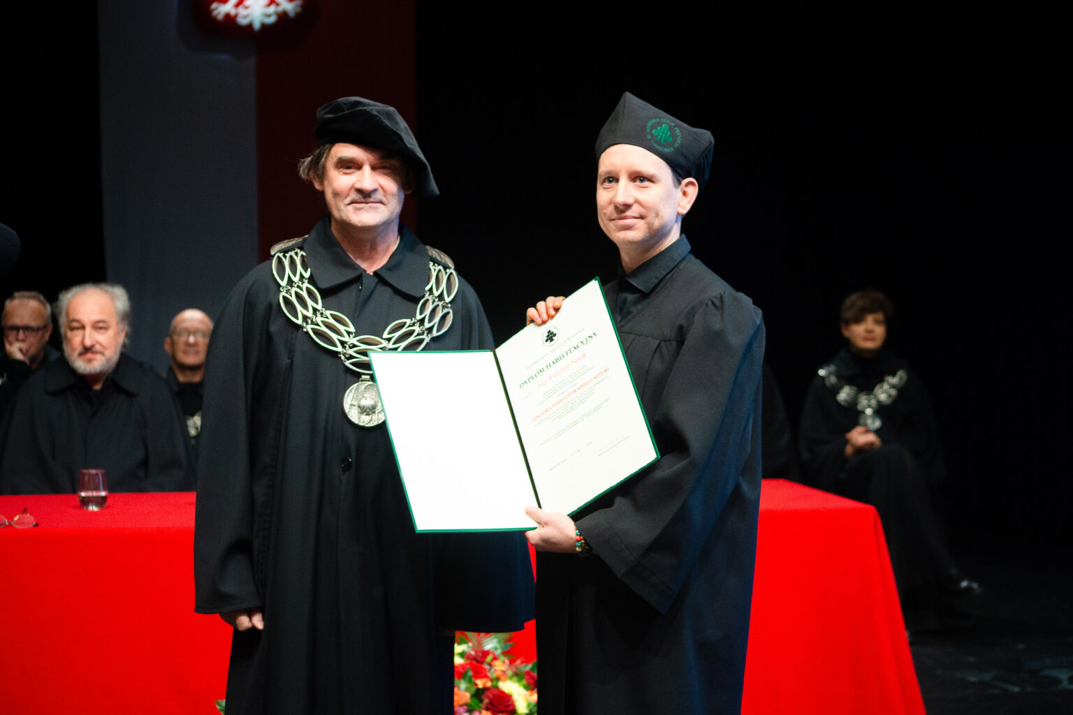 dwaj mężczyźni w strojach reprezentacyjnym ASP w Warszawie, jeden z nich prezentuje dyplom