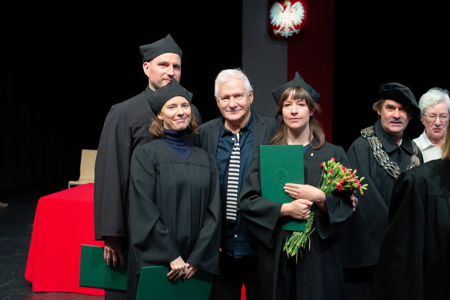 trzy osoby w strojach reprezentacyjnym ASP w Warszawie oraz mężczyzna w czarnym garniturzw i krawacie w biało-czarne pasy, jedna z kobiet trzyma dyplom i kwiaty