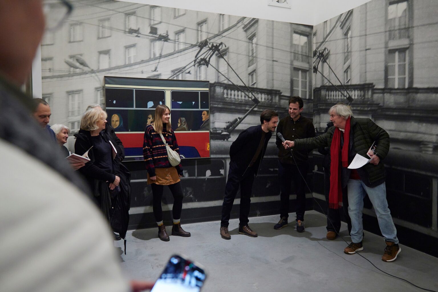 pięć osób stoi na tle fototapety z czarno-białym zdjęciem miasta i kolorowy obrazem przedstawiającym ludzi w czerwonym autobusie
