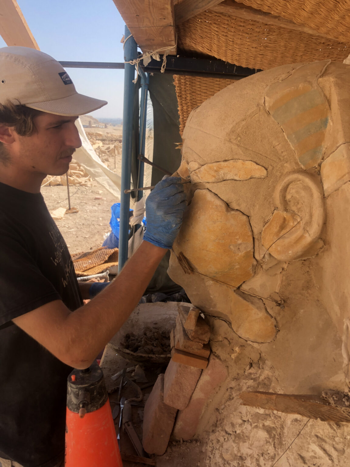 Chłopak w czapeczce z daszkiem, w rękawiczkach wykonuje prace konserwatorskie przy rzeźbie sfinksa