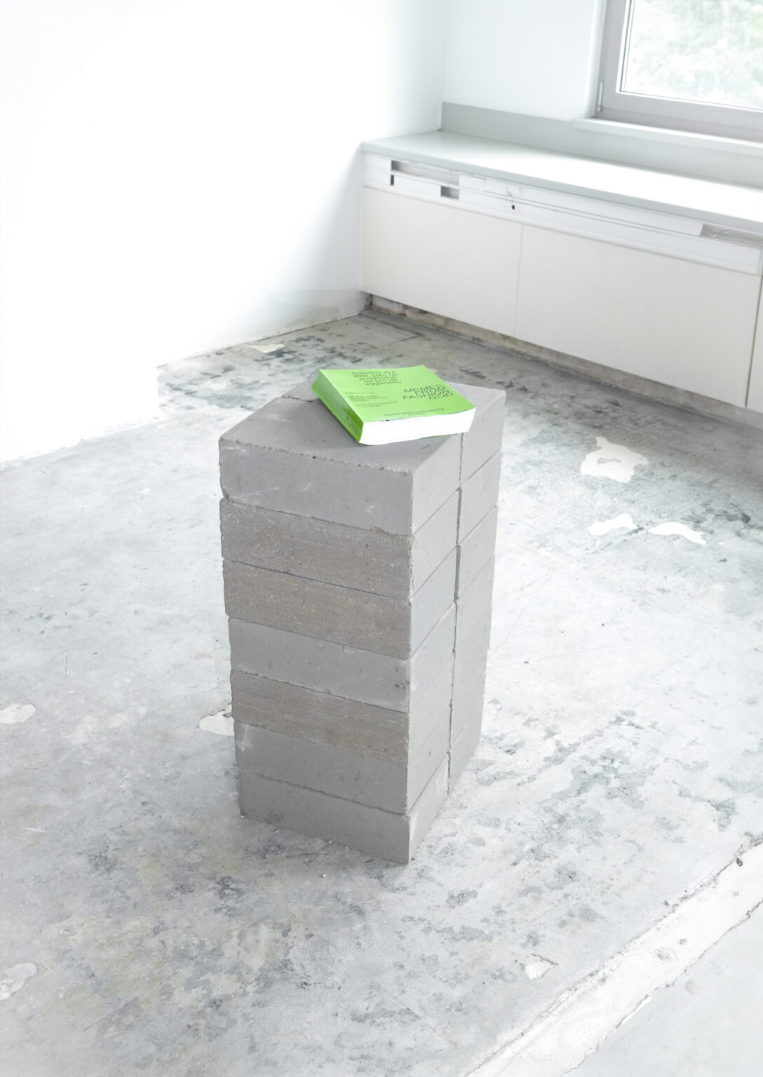 szare cegły ustawione jedna na drugiej, na górze książka z jasnozieloną okładką, betonowe wnętrze wokółj