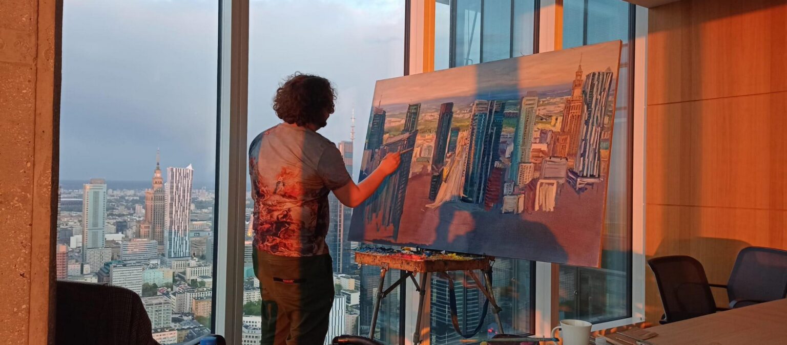 zdjęcie mężczyzny z pędzlem w ręku, stojącego przy sztaludze z obrazem, za oknem panorama miasta