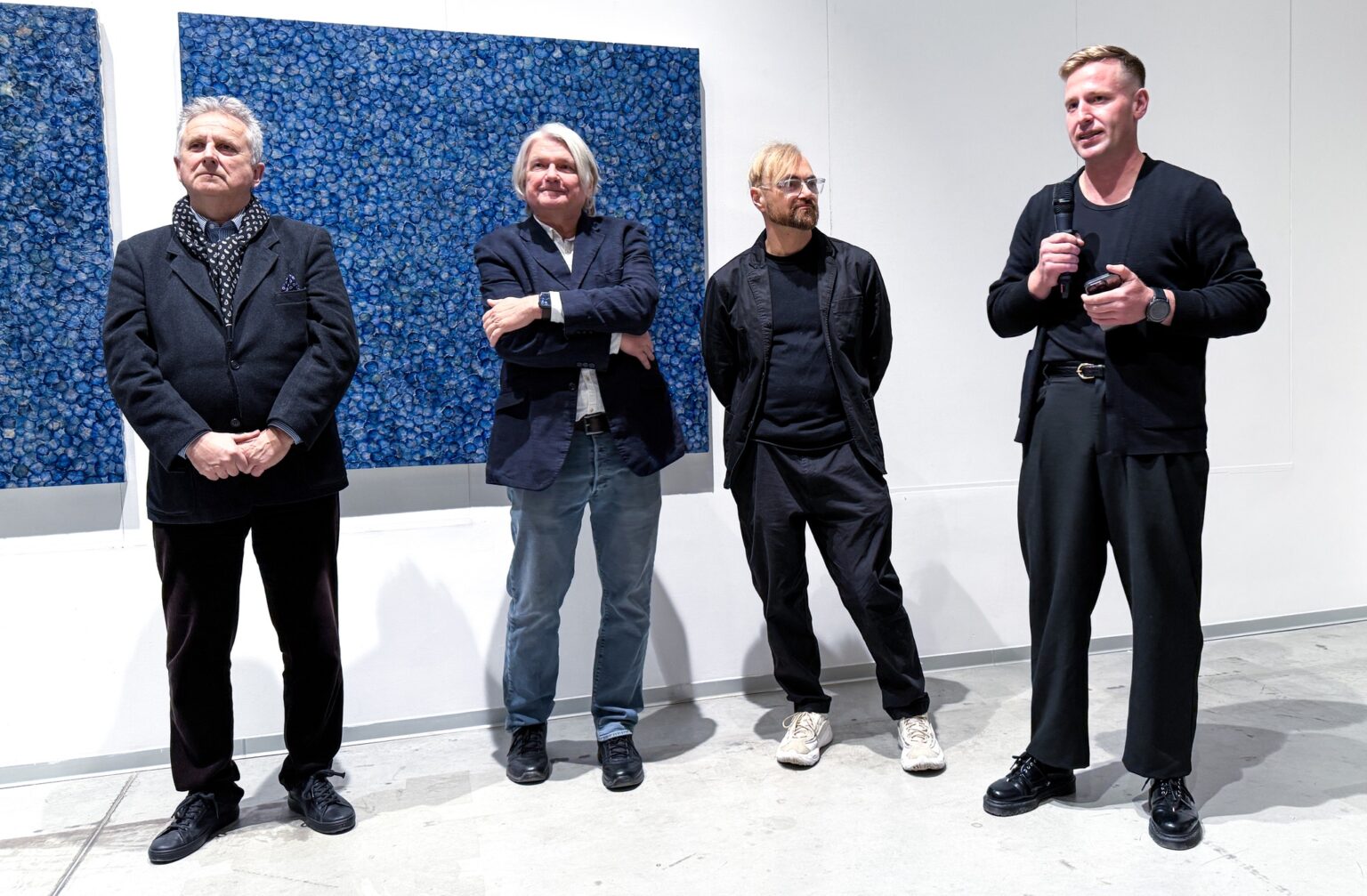 czterech mężczyzn, jeden mówi do mikrofonu, w tle dużych rozmiarów obraz abstrakcyjny