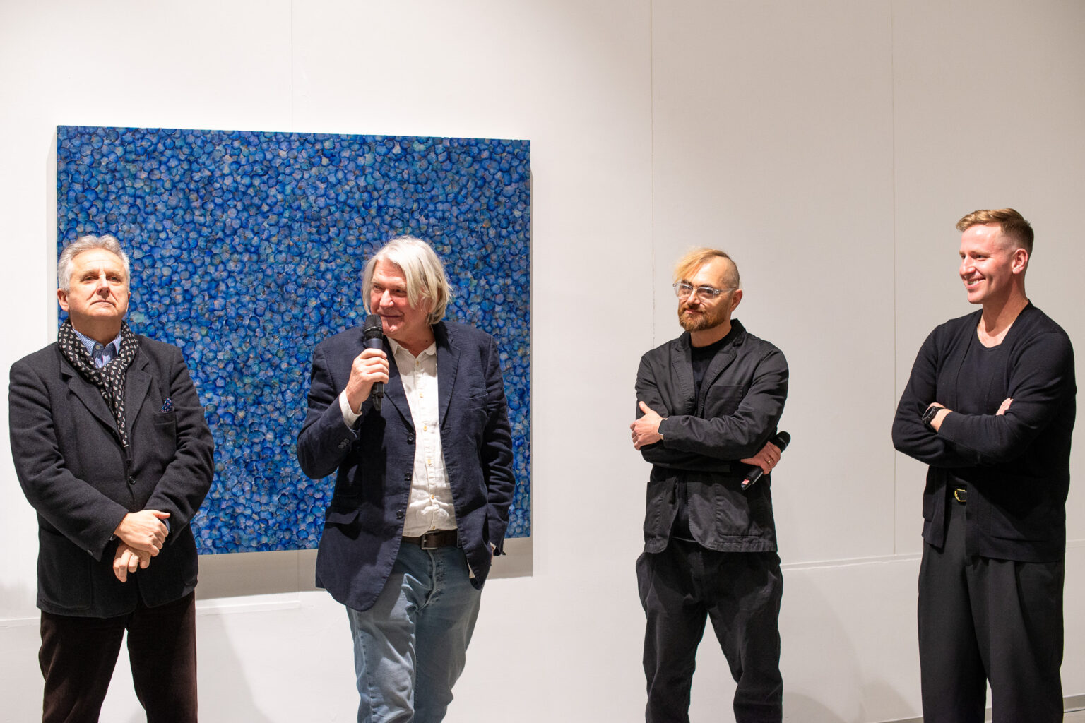 czterech mężczyzn, jeden mówi do mikrofonu, w tle dużych rozmiarów obraz abstrakcyjny