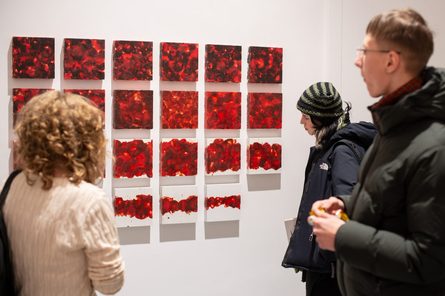 trzy osoby przyglądają się serii niewielkich obrazów w kolorze czerwonym