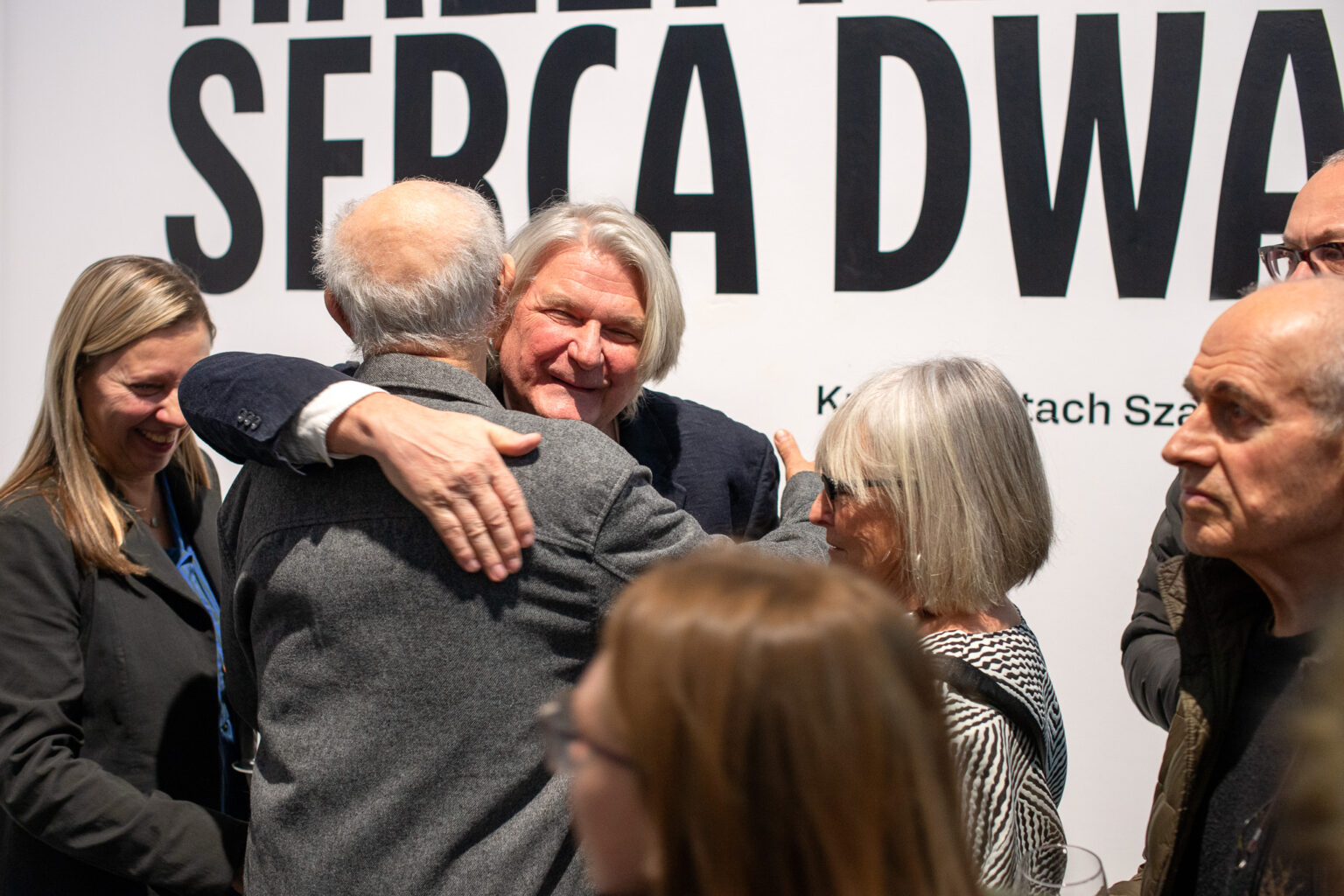 kilka osób, dwaj mężczyźni obejmują się, w tle fragment tytułu wystawy