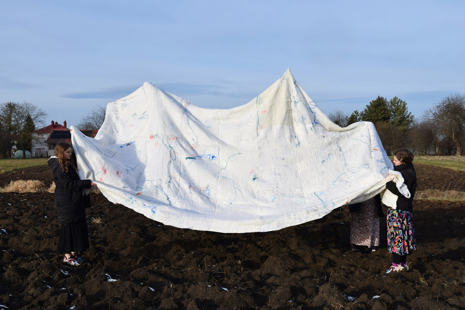 dwie osoby w polu trzymają w wyciągniętych rękach dużą białą płachtę ze śladami farb; na tle niebieskiego nieba