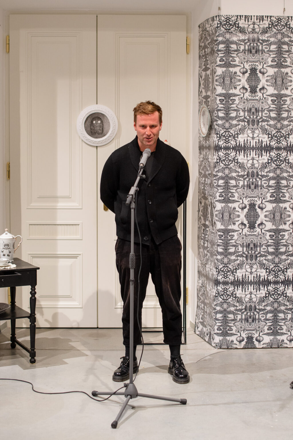 mężczyzna ubrany na czarno przy mikrofonie, wystawa w tle