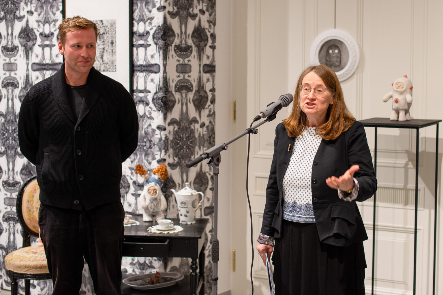 kobieta przy mikrofonie, obok mężczyzna ubrany na czarno, wystawa w tle