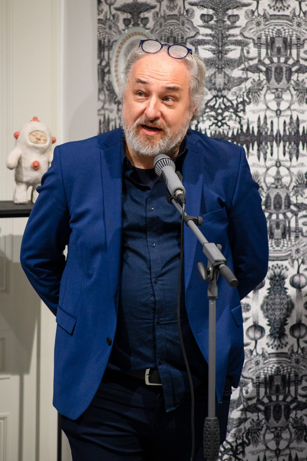 mężczyzna z brodą w niebieskiej marynarce przy mikrofonie, w tle wystawa