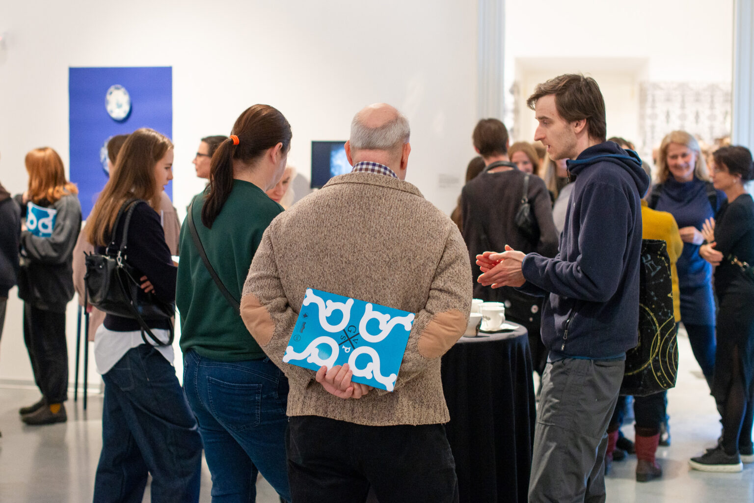 grupa osób zwiedzających w przestrzeni wystawy, mężczyzna tyłem, za plecami trzyma folder wystawy
