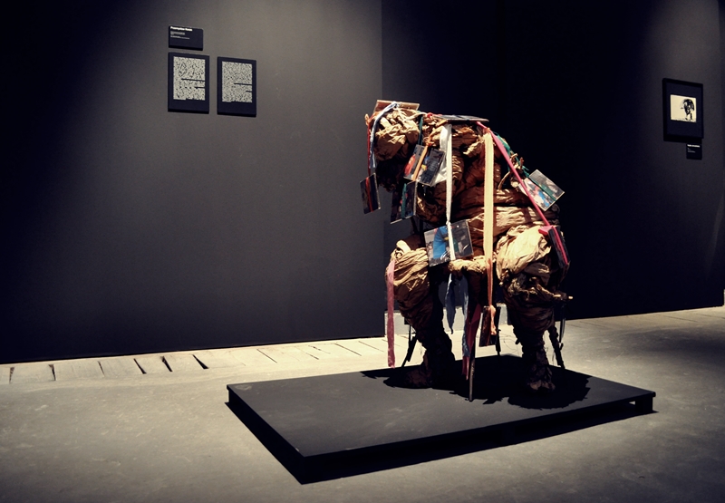 Widok na salę w galerii sztuki. Na tle czarnych ścian na środku sali stoi niski, czarny postument, na którym stoi rzeźba wykonana ze szmat i materiałów. Rzeźba kształtem przypomina siedzącą, pochyloną do przodu figurę ludzką. Z góry rzeźby zwisają wstążki, do których przyczepione są nieduże zdjęcia.