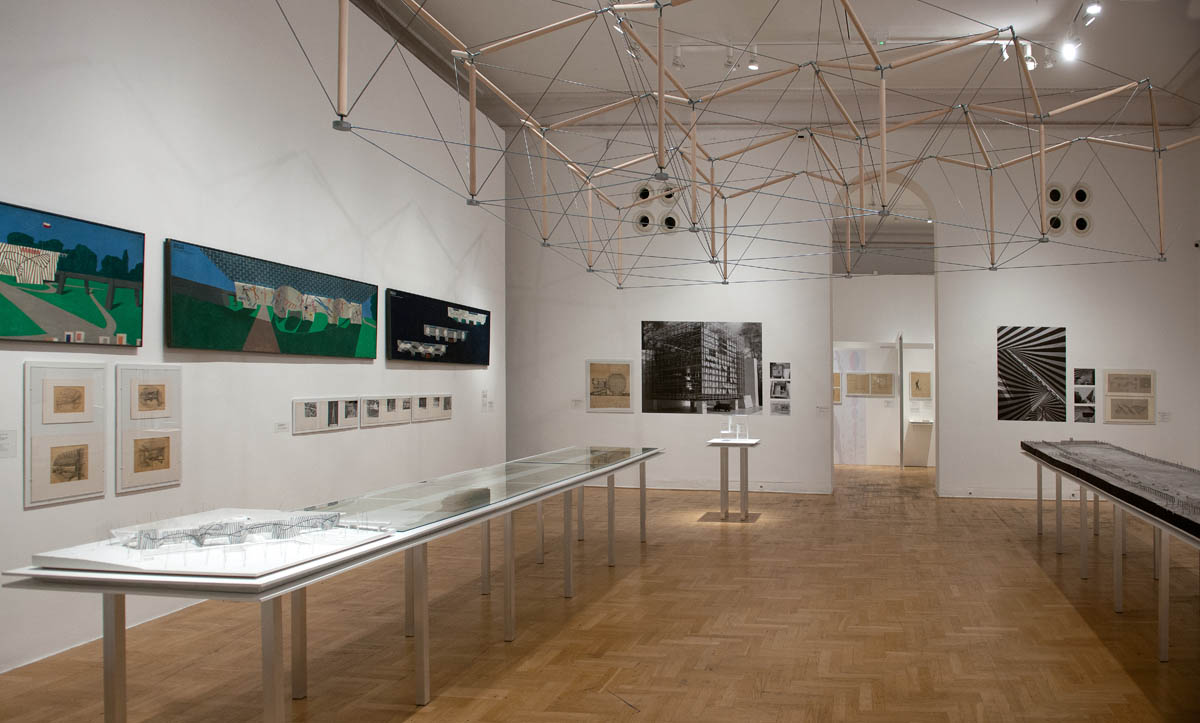 Wnętrze galerii sztuki z wystawą architektoniczną. Na ścianach wiszą kolorowe obrazy i czarno-białe fotografie przedstawiające nowoczesne budynki i konstrukcje. W centralnej części sali znajdują się szklane gabloty z makietami architektonicznymi oraz instalacja z drewnianych prętów i linek zawieszona pod sufitem. Jasne ściany, drewniana podłoga i punktowe oświetlenie podkreślają estetyczny i nowoczesny charakter przestrzeni.