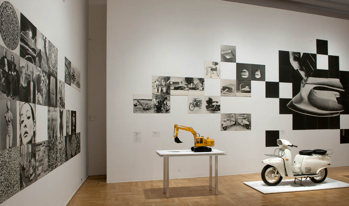 Wnętrze galerii sztuki z ekspozycją poświęconą wzornictwu przemysłowemu i fotografii. Na ścianach umieszczono kolaże czarno-białych zdjęć, przedstawiających ludzi, przedmioty codziennego użytku i elementy architektoniczne. Na pierwszym planie widoczna jest żółta miniaturowa koparka ustawiona na białym stole oraz biały skuter na podświetlonym podeście. Wystawa łączy sztukę wizualną z prezentacją wzornictwa użytkowego, tworząc spójną narrację o designie i kulturze przemysłowej.