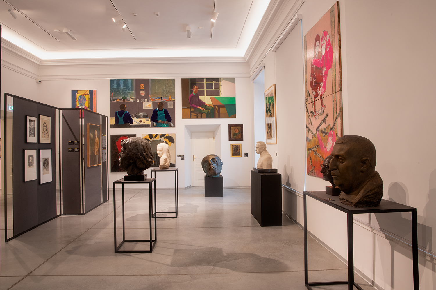 Wnętrze galerii sztuki z ekspozycją obejmującą rzeźby i malarstwo. W centralnej części sali znajdują się popiersia i głowy rzeźbiarskie wykonane z różnych materiałów, umieszczone na minimalistycznych metalowych i drewnianych postumentach. W tle na ścianach wiszą obrazy w różnych stylach – od realistycznych po ekspresyjne i abstrakcyjne, ukazujące sceny z postaciami, portrety oraz kompozycje figuratywne. Po lewej stronie widoczne są przegrody ekspozycyjne z grafikami i rysunkami.