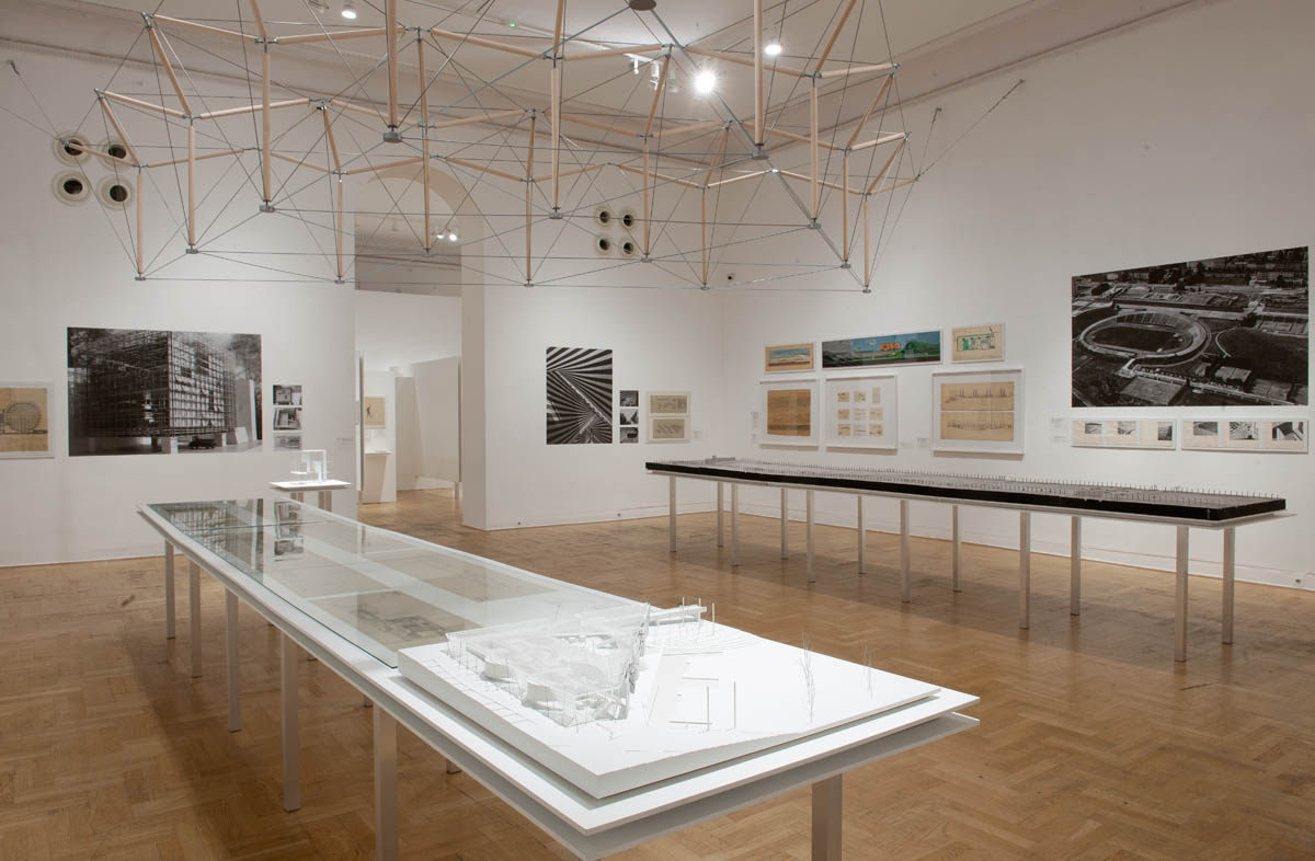 Wnętrze nowoczesnej galerii sztuki z wystawą poświęconą architekturze i urbanistyce. W centrum sali znajdują się długie stoły z makietami architektonicznymi, osłoniętymi szklanymi gablotami. Na ścianach rozmieszczone są czarno-białe fotografie budynków, plany architektoniczne oraz kolorowe rysunki projektowe. Nad przestrzenią wystawową zawieszona jest geometryczna instalacja z drewnianych prętów i napiętych linek, nadająca wnętrzu nowoczesny charakter.