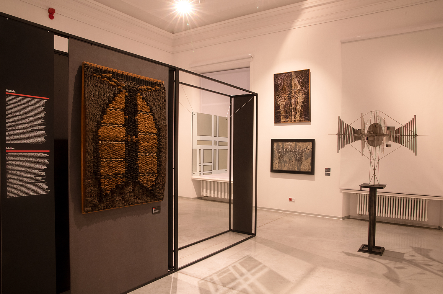 Wnętrze galerii sztuki, na pierwszym planie po lewej stronie znajduje się duża tkanina artystyczna wykonana z naturalnych materiałów, o ciepłych brązowych i czarnych tonach. Po prawej stronie widoczna jest metalowa rzeźba o geometrycznej, symetrycznej konstrukcji, przypominająca futurystyczną strukturę. Na ścianach zawieszone są abstrakcyjne obrazy w ciemnej kolorystyce. Wystawa prezentowana jest w nowoczesnej, jasnej przestrzeni z minimalistycznym designem i subtelnym oświetleniem.