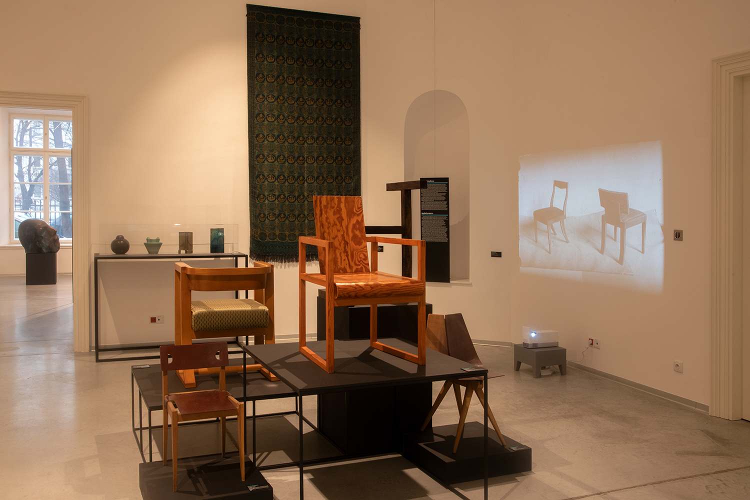 Wystawa poświęcona projektowaniu mebli i wzornictwu. W centralnej części ekspozycji znajduje się kilka drewnianych krzeseł o różnorodnych formach i wykończeniach, ustawionych na metalowych podestach. W tle na ścianie zawieszono dekoracyjną tkaninę o ciemnozielonym wzorze. Po prawej stronie wyświetlana jest projekcja z rzutnika. W głębi sali widać przeszkloną gablotę z ceramicznymi przedmiotami oraz okno.