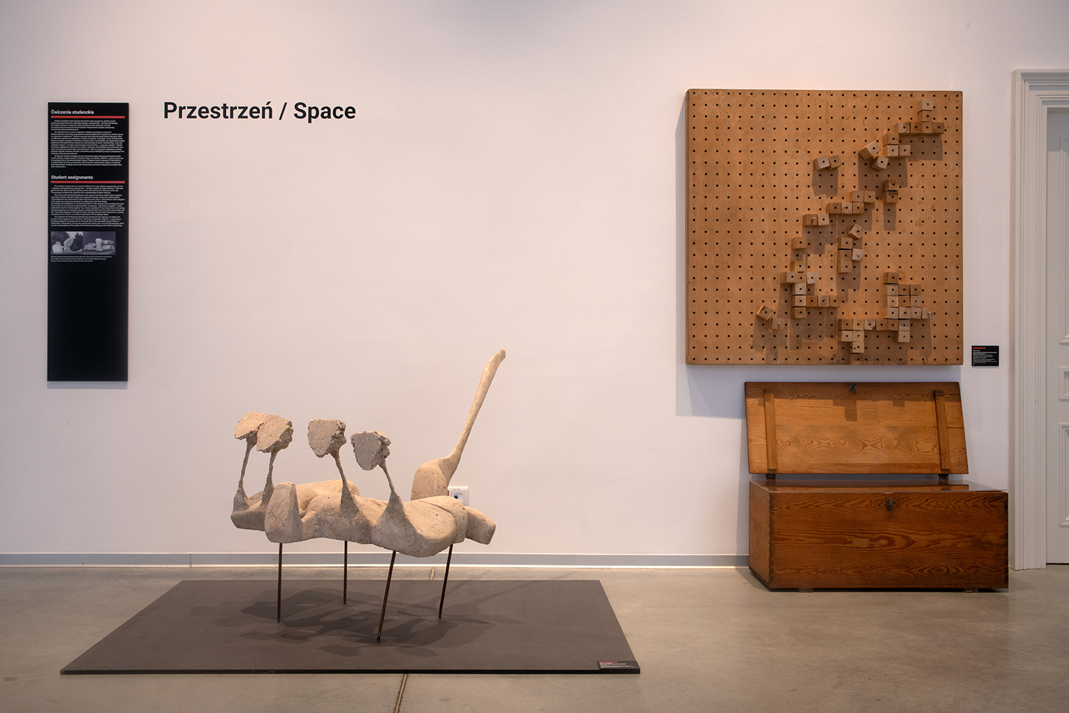 Na zdjęciu widać pomieszczenie galerii. Na pierwszym planie stoi abstrakcyjna rzeźba przypominająca mebel, wykonana z białego gipsu i osadzona na cienkich metalowych nogach. Na białej ścianie wisi drewniana tablica z dziurkami i przyczepionymi do niej drewnianymi kostkami. Pod tablicą stoi otwarta drewniana skrzynia. Po lewej stronie, na ścianie widać napis z czarnych liter: “Przestrzeń / Space”.