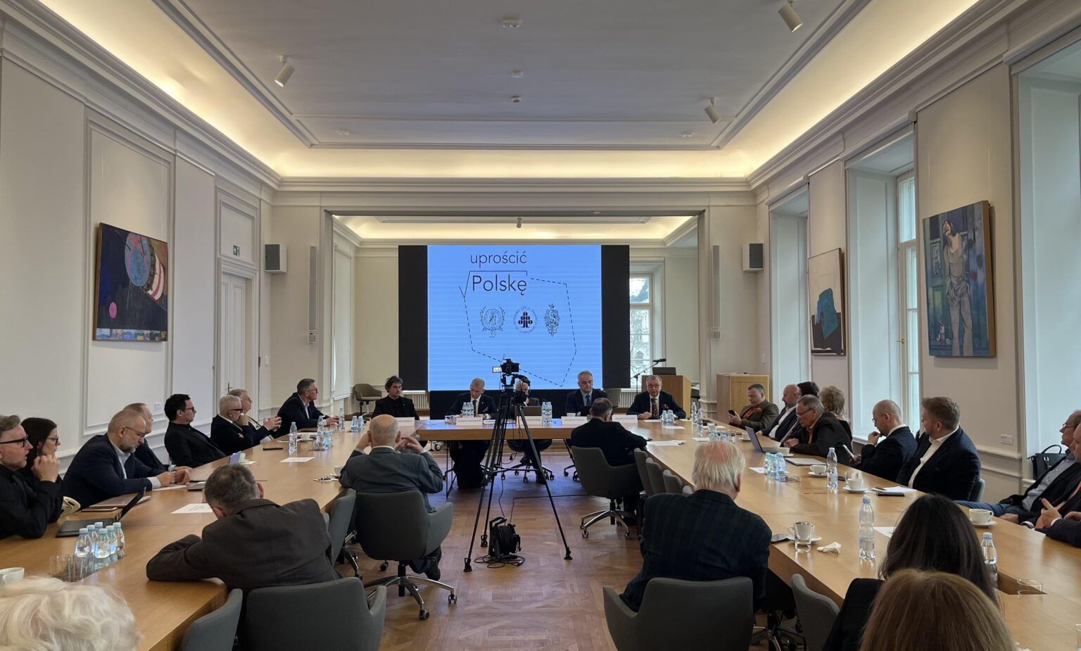 Grupa kilkunastu osób ubranych w garnitury i żakiety siedzi przy stołach ustawionych w literę "U". Przed nimi ekran z wyświetloną grafiką przedstawiającą zarys kształtu Polski z napisem „Uprościć Polskę” i logotypami. Na ścianach wokół nich obrazy.