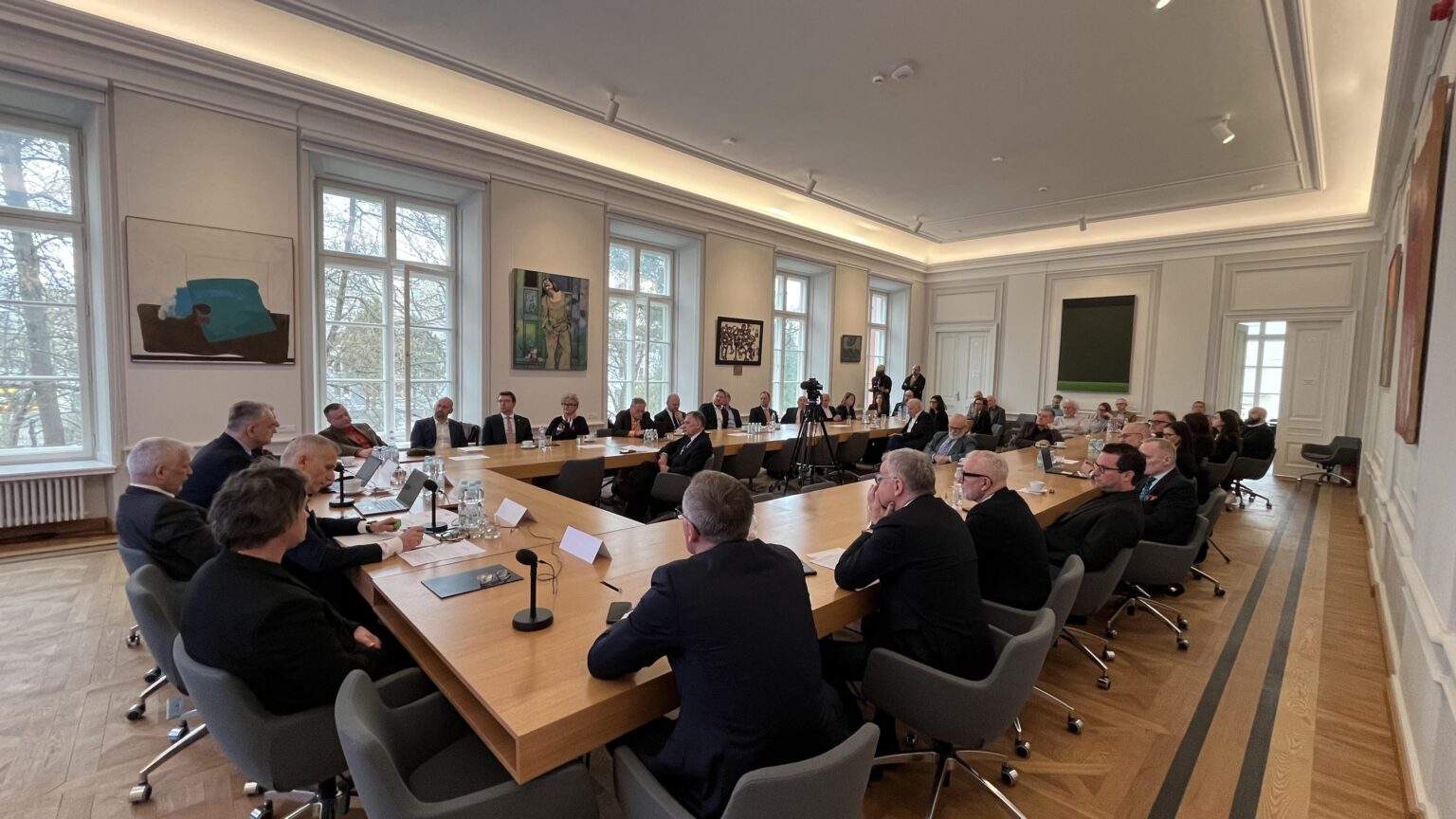 Grupa kilkunastu osób ubranych w garnitury i żakiety siedzi przy stołach ustawionych w literę "U". Przed nimi ekran z wyświetloną grafiką przedstawiającą zarys kształtu Polski z napisem „Uprościć Polskę” i logotypami. Na ścianach wokół nich obrazy.