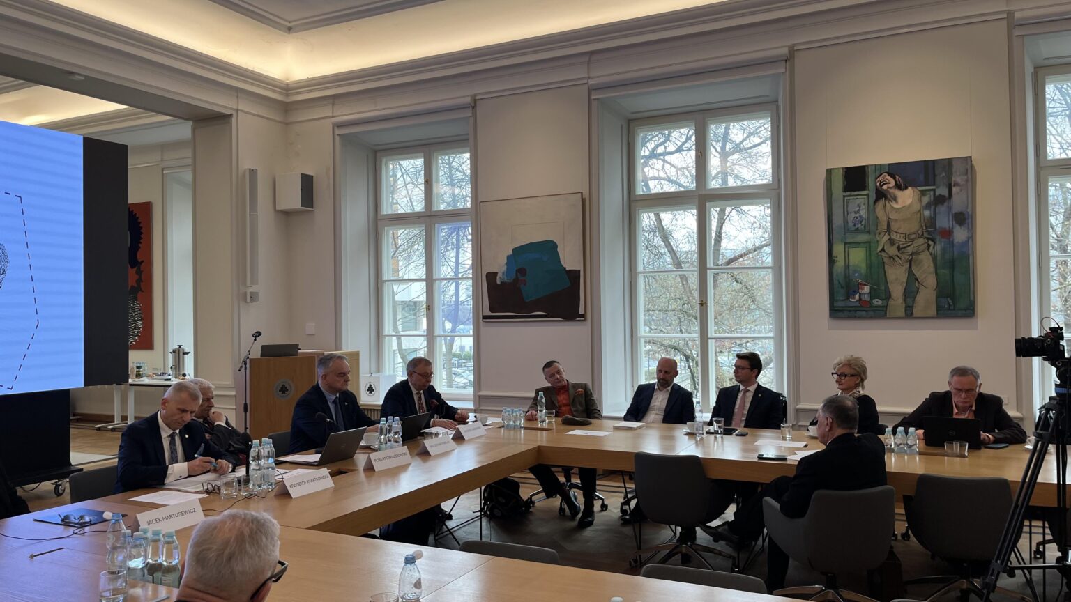 Grupa kilkunastu osób ubranych w garnitury i żakiety siedzi przy stołach ustawionych w literę "U". Przed nimi ekran z wyświetloną grafiką przedstawiającą zarys kształtu Polski z napisem „Uprościć Polskę” i logotypami. Na ścianach wokół nich obrazy.