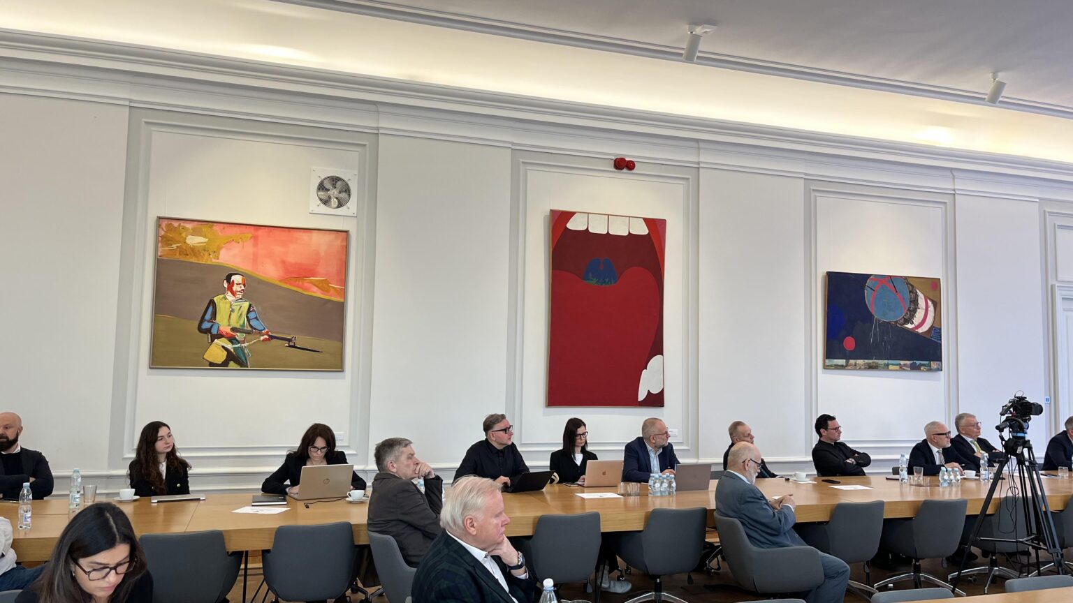 Grupa kilkunastu osób ubranych w garnitury i żakiety siedzi przy stołach ustawionych w literę "U". Przed nimi ekran z wyświetloną grafiką przedstawiającą zarys kształtu Polski z napisem „Uprościć Polskę” i logotypami. Na ścianach wokół nich obrazy.