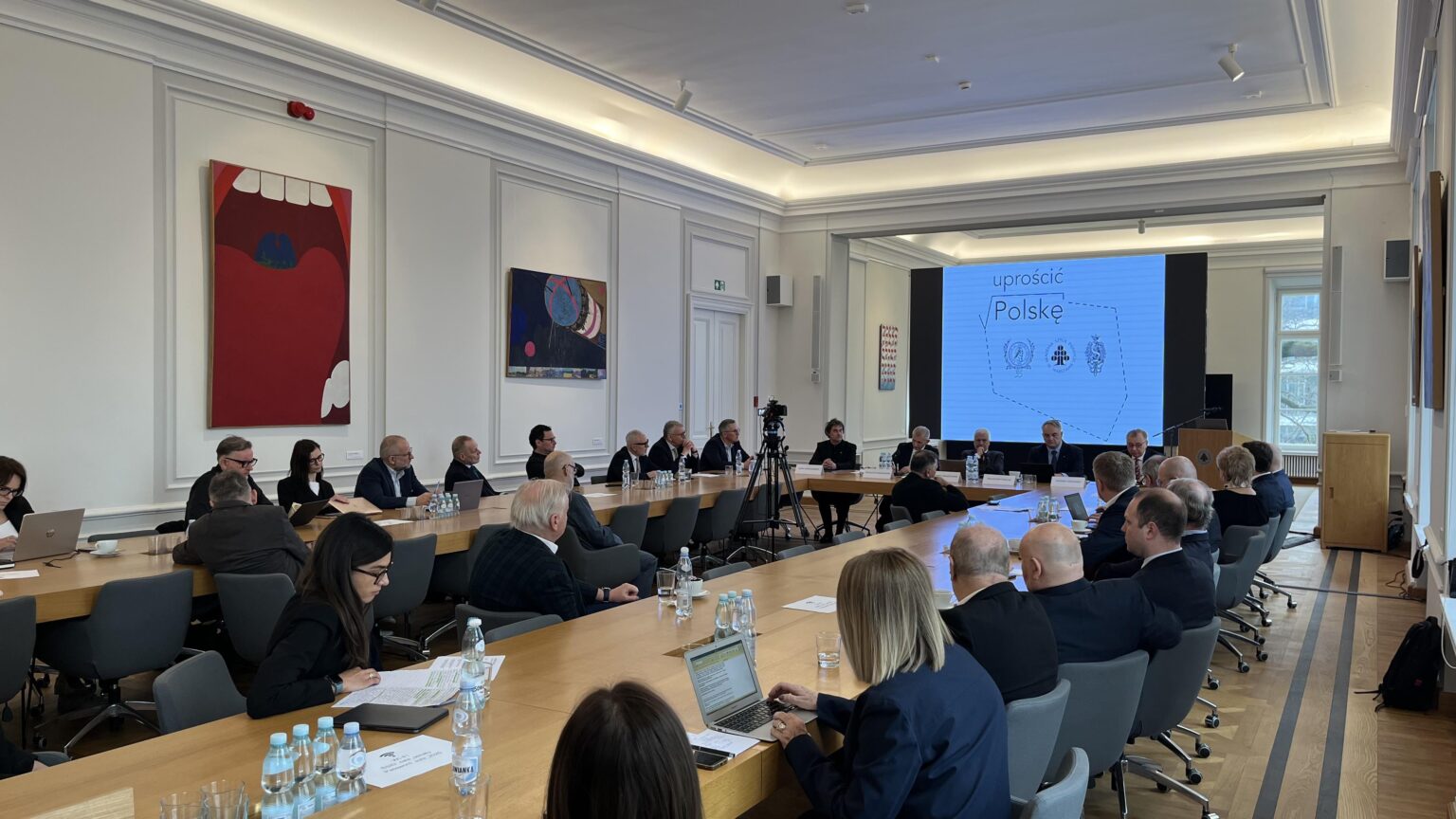 Grupa kilkunastu osób ubranych w garnitury i żakiety siedzi przy stołach ustawionych w literę "U". Przed nimi ekran z wyświetloną grafiką przedstawiającą zarys kształtu Polski z napisem „Uprościć Polskę” i logotypami. Na ścianach wokół nich obrazy.
