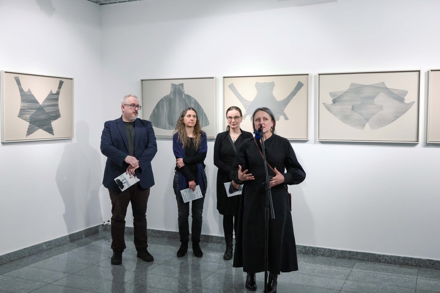 cztery osoby w galerii, kobieta mówi do mikrofonu, rysunki na ścianie w tle