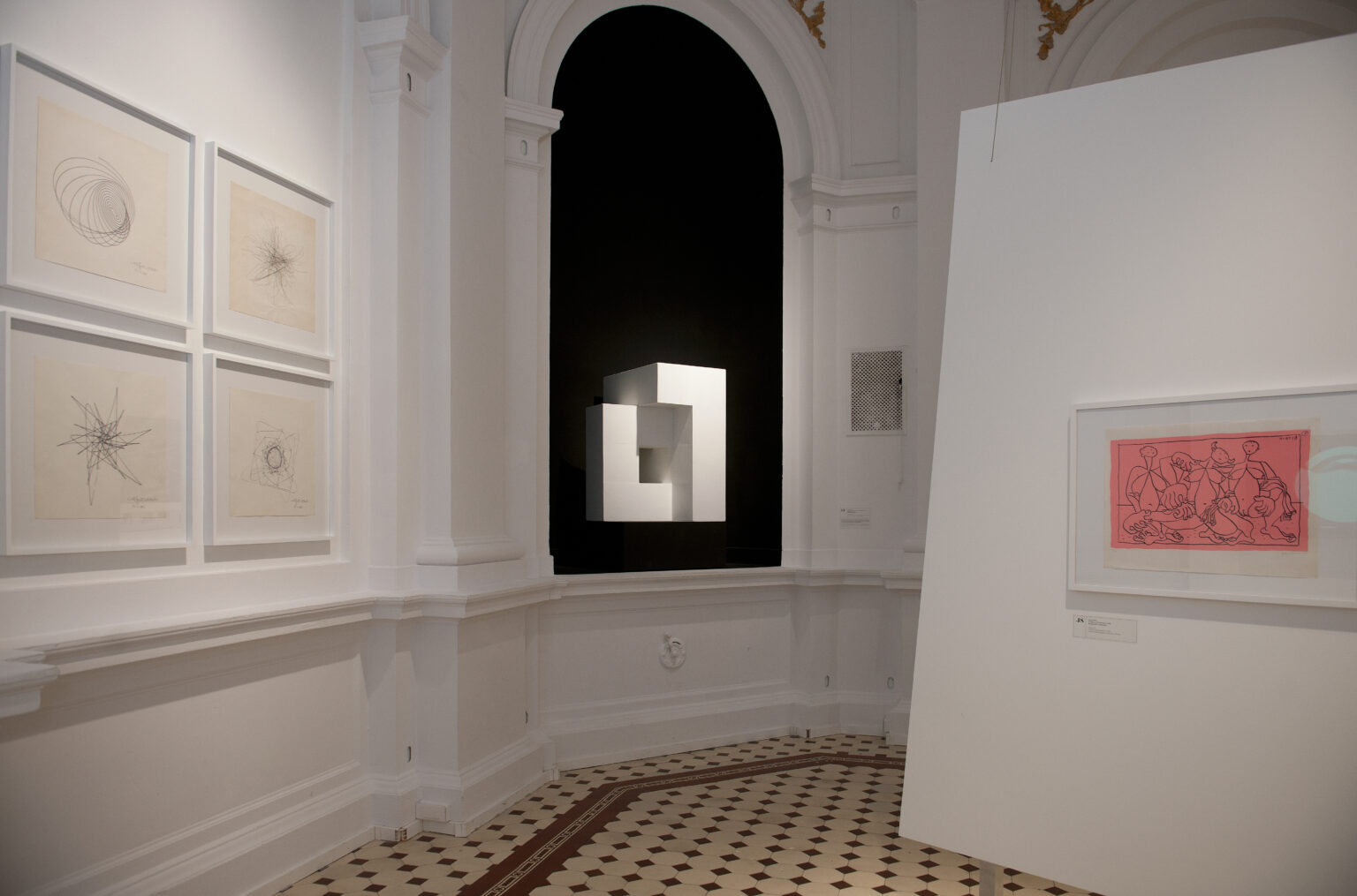 Pomieszczenie w galerii sztuki. W białych ramkach, na tle białej ściany wiszą oprawione abstrakcyjne rysunki. W centrum zdjęcia znajduje się czarna wnęka, w której wisi geometryczna, przestrzenna figura. Po prawej stronie zdjęcia, znajduje się biała ściana działowa, w centrum wisi rysunek przedstawiający postacie na różowym tle.