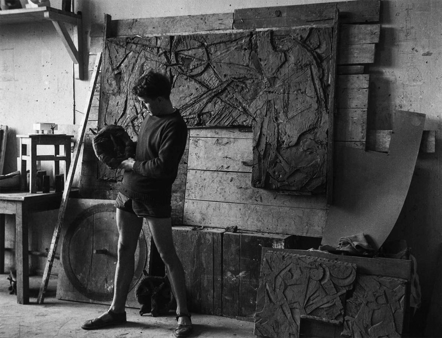 czarno-białe zdjęcie: mężczyzna stoi w pracowni przyglądając się okrągłemu obiektowi w dłoniach