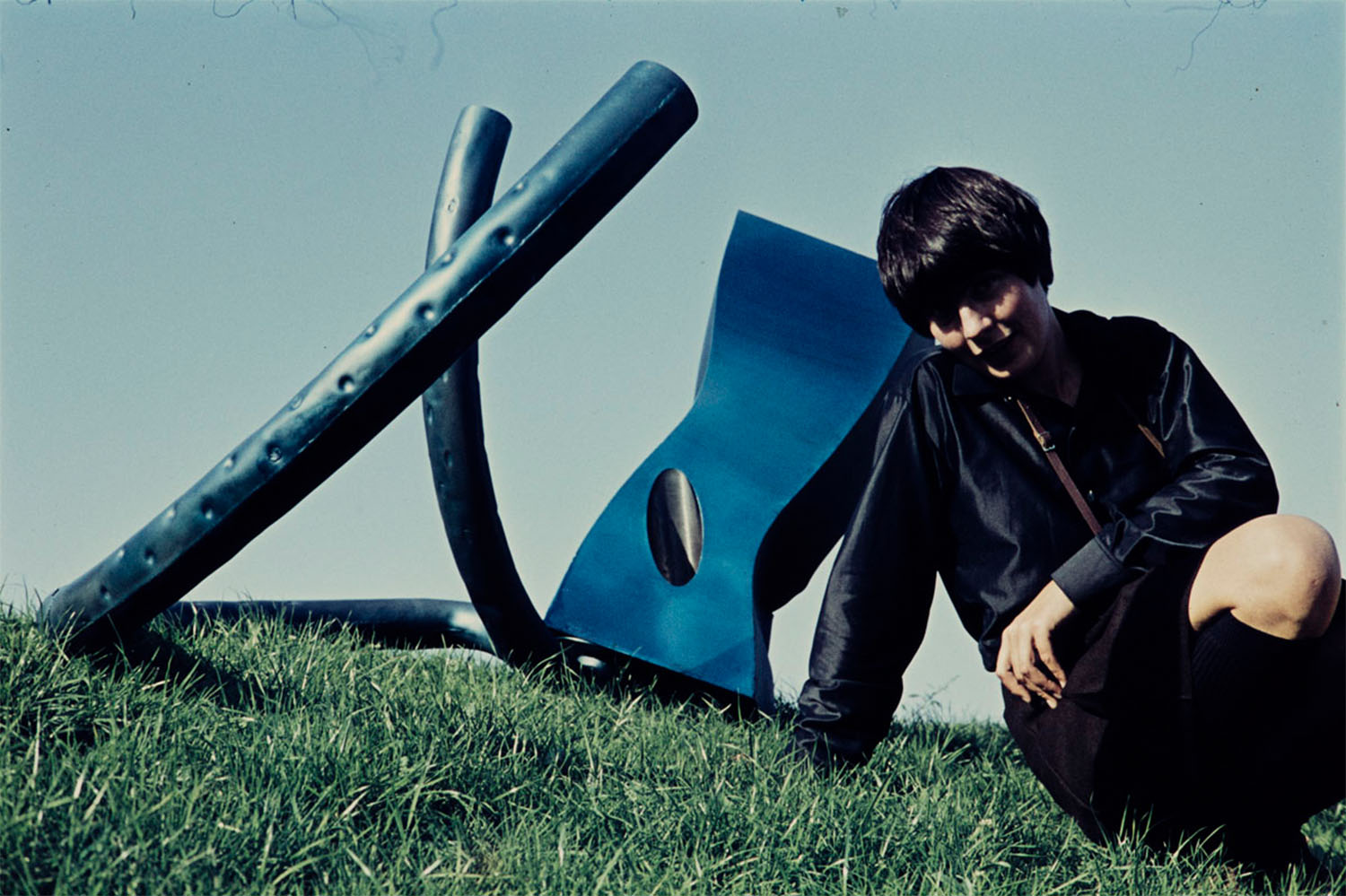 kolorowe zdjęcie: krótkowłosa kobieta siedzi na trawie obok dużej metalowej rzeźby o abstrakcyjnym kształcie