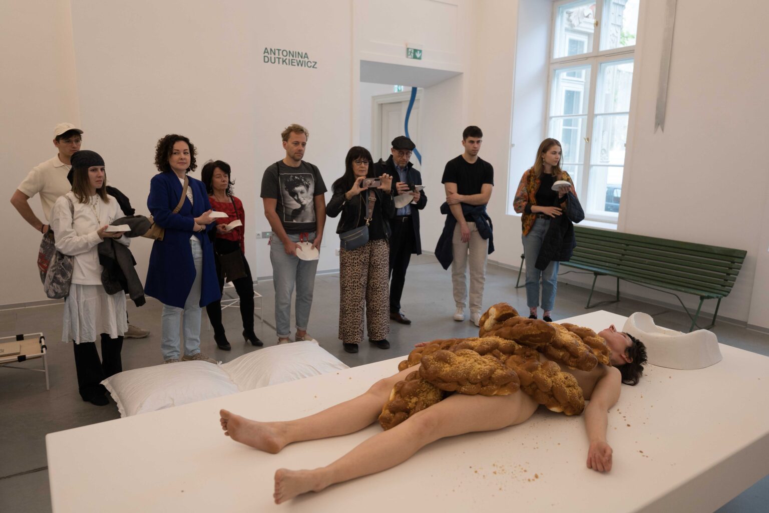 grupa osób stoi trzymając w rękach białe naczynia, na pierwszym planie naga kobieta leży na białym stole, na niej sterta chałek