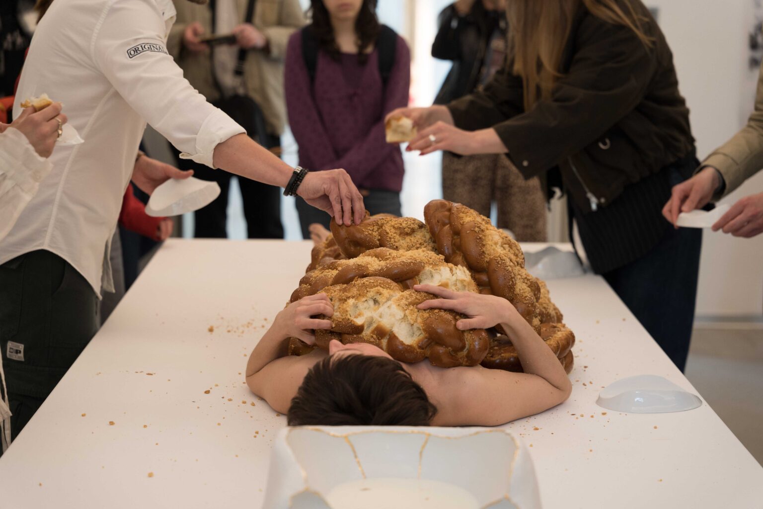 na pierwszym planie naga kobieta leży na białym stole, na niej sterta chałek, wokół ludzie odrywający kawałki chałki