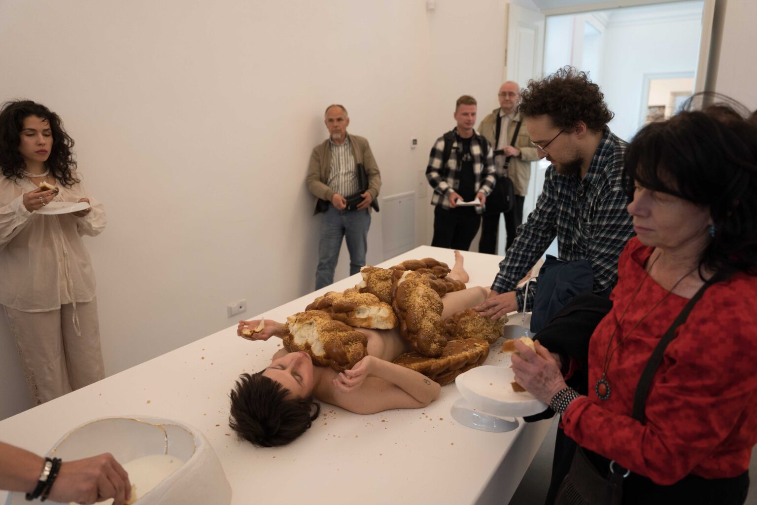 naga kobieta leży na białym stole, na niej sterta chałek, wokół ludzie odrywający kawałki chałki