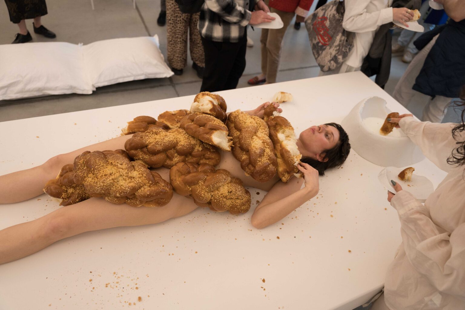 naga kobieta leży na białym stole, na niej sterta chałek, wokół ludzie